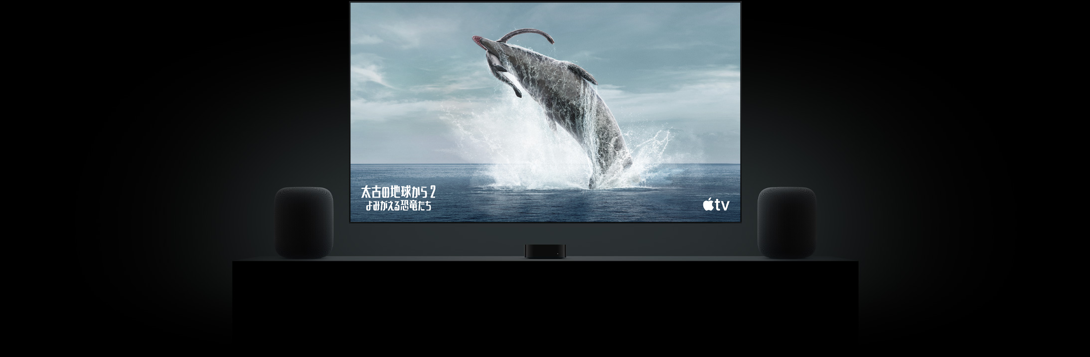 フラットテレビの大きなスクリーン上に「太古の地球から?よみがえる恐竜たち?」の恐竜の鮮明な映像が映し出されている。リビングルームのキャビネットの上に置かれた2臺(tái)のHomePodスピーカー。その間にApple TVが配置され、その上にテレビが掛けられている