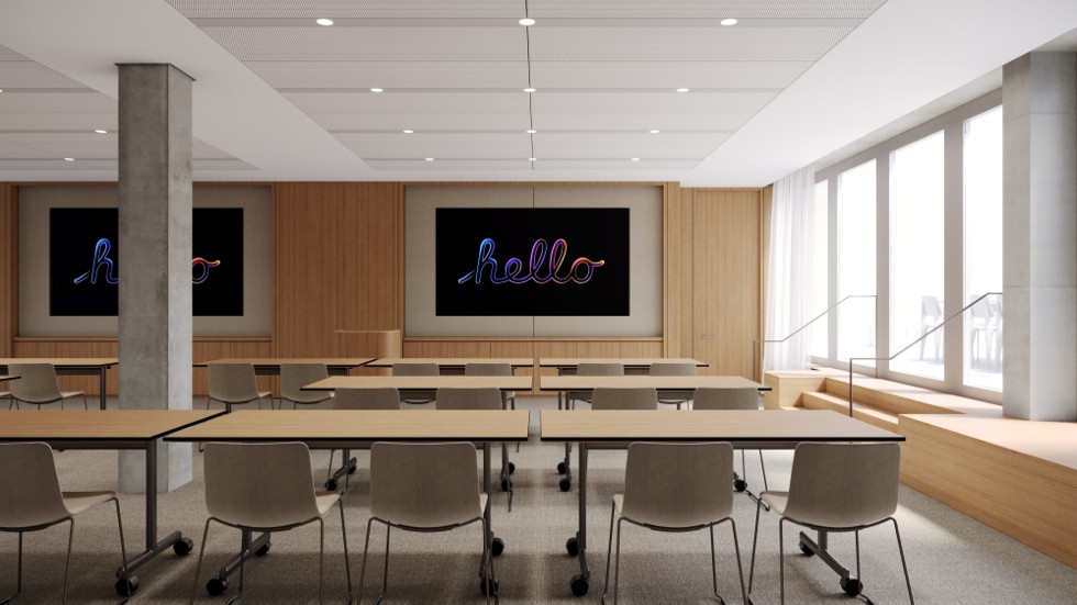 An empty training room is set up with tables and chairs facing two large screens on a wood-paneled wall, each displaying the word “hello” in a cursive neon script.