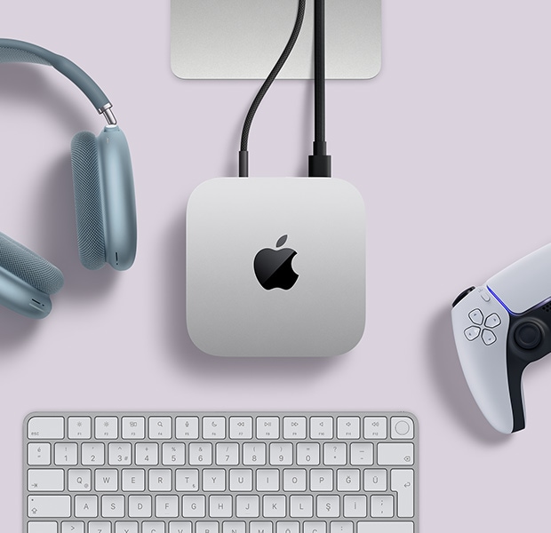 Arka ba?lant? noktalar?na iki adet kablonun ba?l? oldu?u gümü? rengi Mac mini ile yan?nda yer alan mavi bir AirPods Max, gümü? rengi bir Magic Keyboard ve kablosuz bir oyun kumandas?n?n üstten g?rünümü