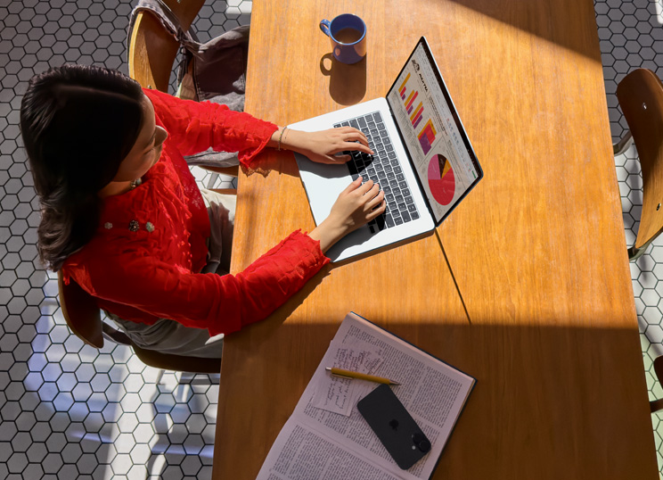 Iemand die een MacBook gebruikt aan een tafel, met een mok, smartphone en papieren in de buurt.