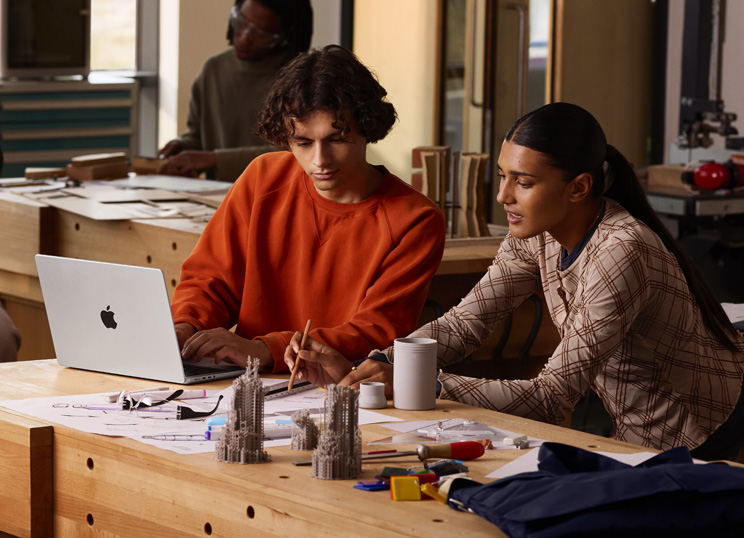 Duas pessoas colaborando em um projeto à mesa: uma digitando em um MacBook, a outra usando um lápis e uma régua no papel, cercadas por materiais de arte