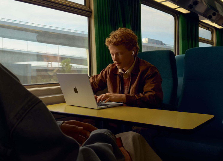 Pessoa em um trem, sentada junto à janela, usando AirPods e trabalhando em um MacBook