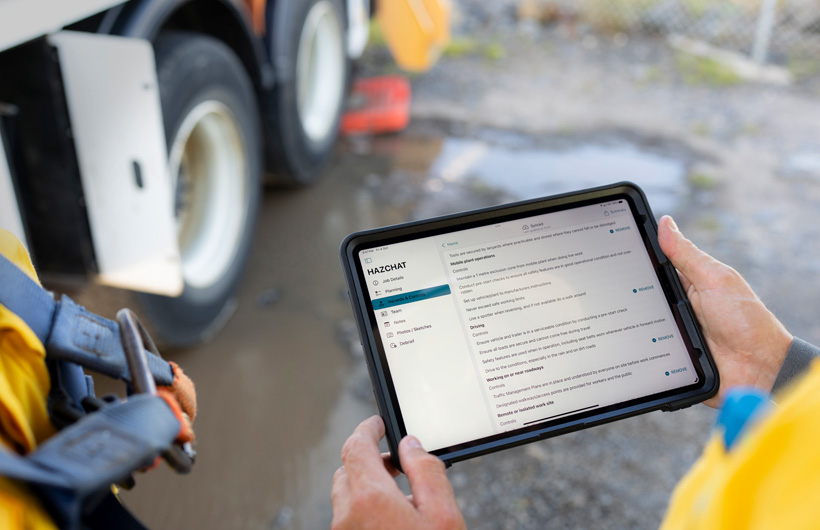 Hender holder en iPad med HazChat-appen ?pen, og p? skjermen vises Job Details, Planning, Hazards og Controls. I bakgrunnen st?r en person ved en s?ledam p? arbeidsstedet.