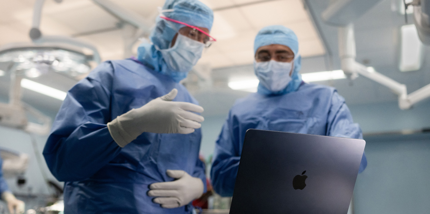Zwei Chirurgen im Operationssaal schauen auf ein MacBook, wobei einer eine Geste mit der Hand macht, w?hrend der andere beobachtet